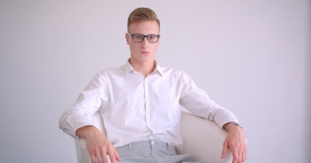 Портрет крупным планом молодого красивого кавказского бизнесмена в очках, сидящего в кресле и счастливо смотрящего на камеру в белой комнате, улыбающегося в помещении — стоковое видео