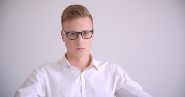 Крупный план портрета молодого привлекательного кавказского бизнесмена в очках, сидящего в кресле и смотрящего в камеру в белой комнате — стоковое видео