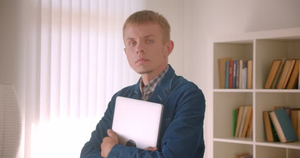 Retrato del bibliotecario caucásico mirando a la cámara con calma sosteniendo el ordenador portátil en las manos sobre los estantes de fondo . — Vídeo de stock