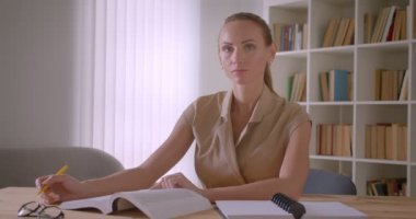 Kütüphanede ciddi ve mütevazı bir şekilde kameraya kamera izleyen kitap ve kağıtlarla çalışan çekici öğretmen.