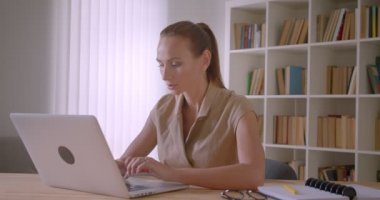 Dizüstü bilgisayarda yazan zarif beyaz iş kadını meşgul ken kameraya döner ve ofiste gülümsüyor.