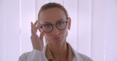 Beyaz önlüklü başarılı beyaz kadın doktor gözlüklerini düzeltip hastanede kameraya gülümsüyor.