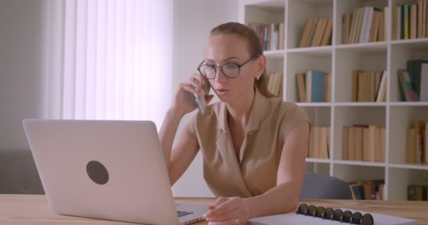 Seriöse kaukasische Geschäftsfrau mit Brille, die auf dem Handy spricht, schaut aufmerksam in Laptop im Büro. — Stockvideo