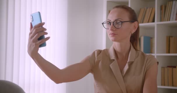 Gözlüklü güzel iş kadını akıllı telefonda selfie yapıyor gülümseyip ofiste mutlu oluyor. — Stok video