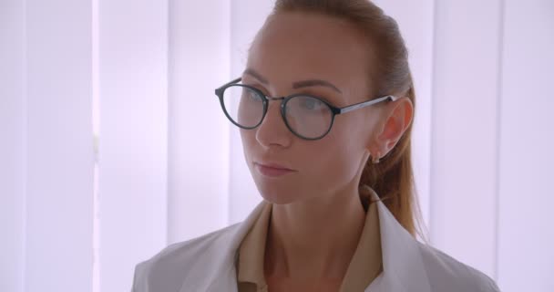 Erfolgreiche kaukasische Ärztin in weißem Mantel und Brille lächelt im Krankenhaus vor Jalousie-Hintergrund in die Kamera. — Stockvideo