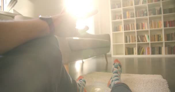 Gros plan de jambes de cameramen assis sur le sol sur le tapis avec des étagères sur le fond avec la lumière du soleil — Video