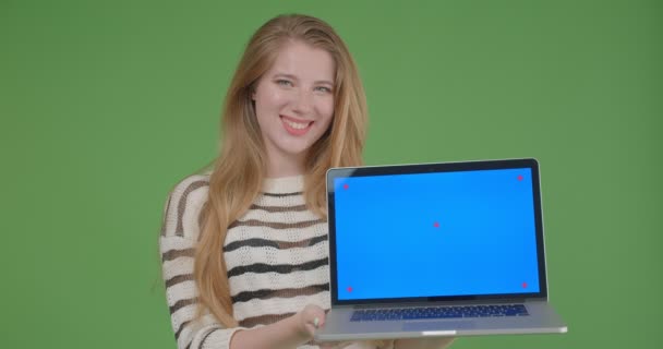 Primer plano de la joven hembra bastante caucásica sosteniendo el portátil y mostrando la pantalla azul a la cámara sonriendo felizmente con el fondo aislado en verde — Vídeos de Stock