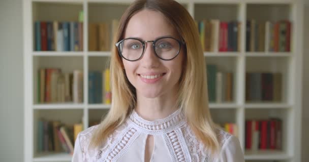 Nahaufnahme Porträt einer jungen hübschen kaukasischen Studentin mit Brille, die fröhlich lächelt und ihr Haar fixiert, während sie in die Kamera in der Universitätsbibliothek blickt — Stockvideo