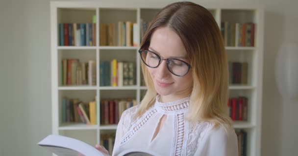 Lähikuva muotokuva nuori kaunis valkoihoinen naispuolinen opiskelija lasit lukeminen kirja hymyillen iloisesti katsot kameraa college kirjastossa — kuvapankkivideo