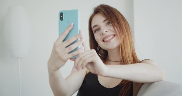 Primer plano de brote de joven seductora hembra caucásica en traje de cuerpo negro tomando selfies en el teléfono sentado en el sofá interior — Vídeos de Stock