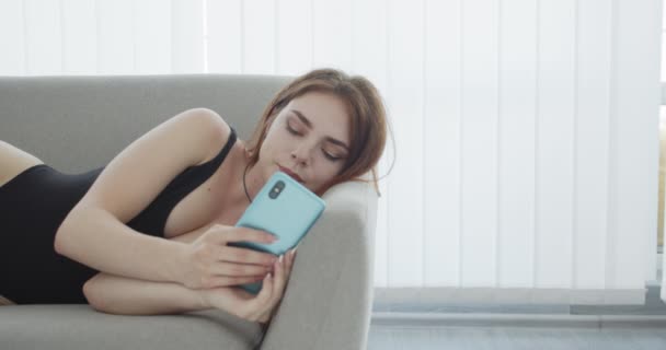 Съемки крупным планом молодой сексуальной белой женщины в черном купальнике, переписывающейся по телефону и расстраивающейся, лежа на диване в помещении — стоковое видео