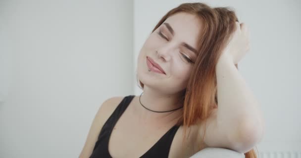 Close-up tiro de jovem sexy caucasiano fêmea no preto bodysuit sorrindo feliz inclinando-se em seu braço sentado no sofá dentro de casa — Vídeo de Stock