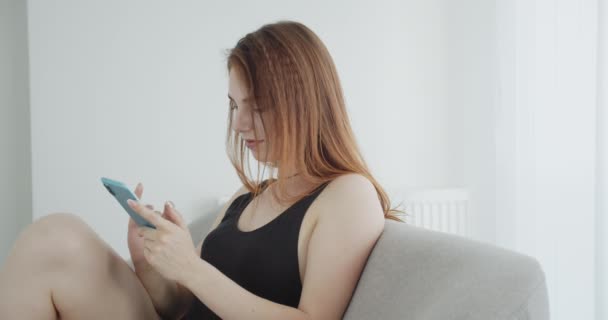 Närbild shoot av unga sexiga kaukasiska kvinna i svart Body med hjälp av telefonen vänder sig till kameran och poserar glatt sitter på soffan inomhus — Stockvideo