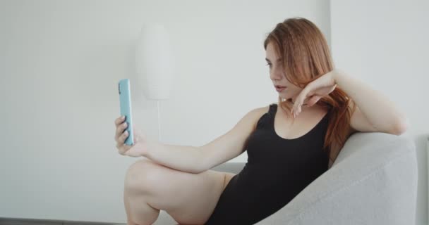 Съемки крупным планом молодой сексуальной белой женщины в черном купальнике, делающей селфи по телефону, сидящей на диване в помещении — стоковое видео