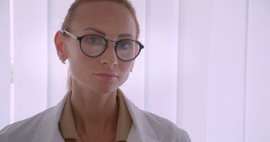 Beyaz ceketli profesyonel beyaz kadın doktor gözlüklerini düzeltir ve hastanede kameraya sakince gülümsüyor.