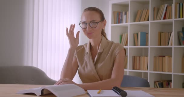Приваблива біла вчителька фіксує окуляри, спокушаючи камеру в бібліотеці . — стокове відео