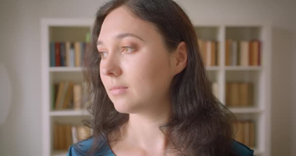 Close-up portret van jonge mooie blanke vrouwelijke student draaien naar camera in de Universiteitsbibliotheek binnen met boekenkasten op de achtergrond — Stockvideo