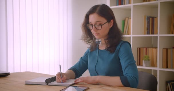 Retrato de cerca de una joven estudiante bastante caucásica en gafas estudiando y escribiendo en el cuaderno usando la tableta en la biblioteca universitaria en el interior — Vídeos de Stock