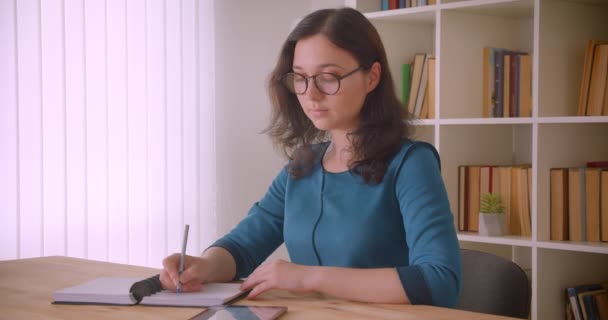 Closeup portrét mladé ženy z kavkazské studentky v brýlích studujících a zapisujících v poznámkovém bloku pomocí tabletu, která se dívá na fotoaparát v univerzitní knihovně uvnitř — Stock video