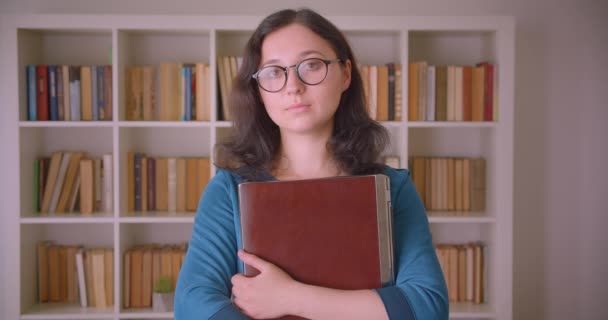 Retrato de cerca de una joven estudiante bastante caucásica con gafas sosteniendo un portátil mirando la cámara de pie en la biblioteca de la universidad en el interior — Vídeos de Stock
