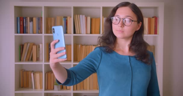 Närbild porträtt av unga ganska kaukasiska kvinnlig student i glasögon tar selfies på telefonen i kollegiet biblioteket inomhus — Stockvideo