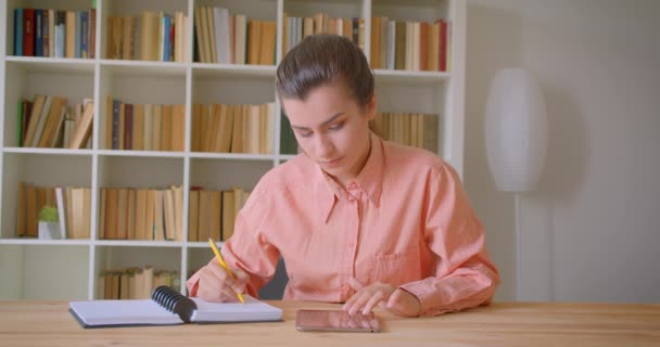 Retrato de cerca de una joven estudiante atractiva que estudia y usa la tableta mirando la cámara en la biblioteca de la universidad — Vídeos de Stock