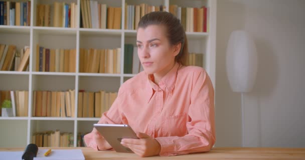 Retrato de cerca de una joven estudiante atractiva que estudia y usa la tableta sentada en la biblioteca de la universidad — Vídeos de Stock