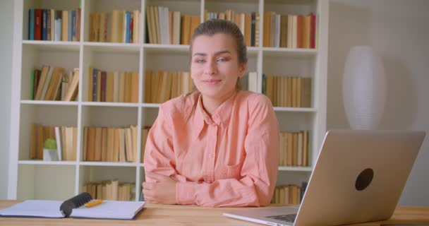 Retrato de cerca de una joven atractiva mujer de negocios sonriendo felizmente mirando a la cámara sentada frente al portátil en la biblioteca dentro — Vídeos de Stock