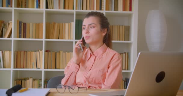 Närbild porträtt av unga attraktiva affärskvinna pratar glatt på telefonen sitter framför den bärbara datorn i biblioteket med bokhyllor på bakgrunden — Stockvideo