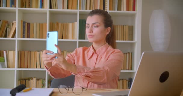 Genç çekici iş kadınının arka planda kitaplıklarla kütüphanede dizüstü bilgisayarın önünde otururken telefonda selfie çektiği yakın çekim portresi — Stok video