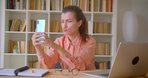 Closeup portrait of young attractive businesswoman taking selfies on the phone posing sitting in front of the laptop with bookshelves on the background — Stock Video