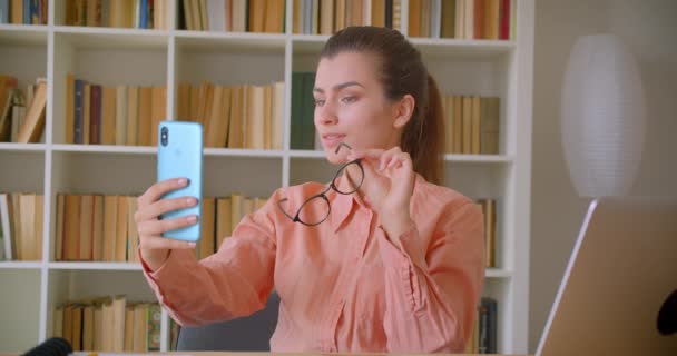 Närbild porträtt av unga attraktiva affärskvinna i glasögon med selfies på telefonen leende glatt sitter framför den bärbara datorn med bokhyllor på bakgrunden — Stockvideo