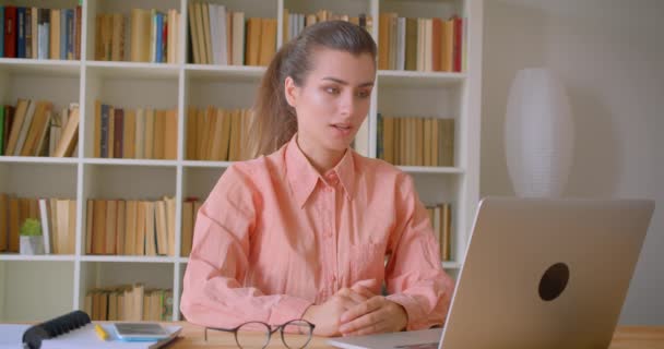 Retrato de close-up de jovem mulher de negócios atraente que tem uma chamada de vídeo formal no laptop na biblioteca dentro de casa — Vídeo de Stock