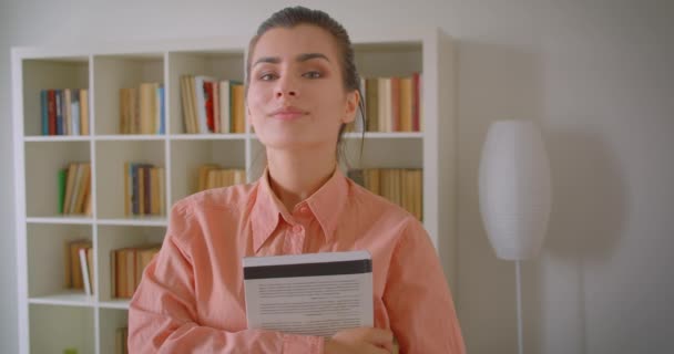 Retrato de cerca de una joven estudiante atractiva mirando a la cámara sonriendo alegremente sosteniendo un libro de pie en el apartamento con estanterías en el fondo — Vídeo de stock