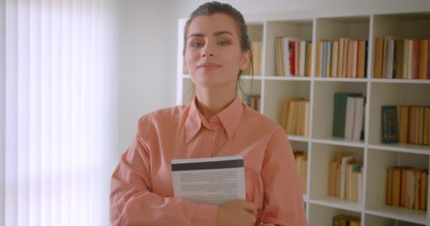 Closeup portréja fiatal vonzó női hallgató nézi kamera mosolygó boldogan tartja a könyvet állt a lakásban könyvespolcok a háttérben — Stock videók