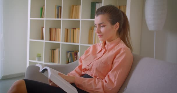 Närbild porträtt av unga attraktiva affärskvinna läsa en bok tittar på kamera leende lyckligt sitter på soffan hemma inomhus — Stockvideo