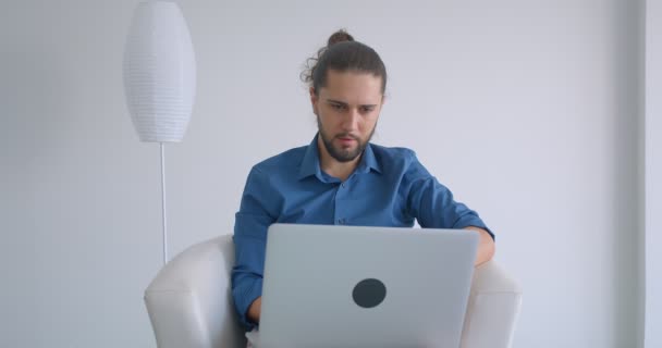 Freelancer progresivo con cola de caballo trabajando con laptop sentado en sillón en cómoda y ligera oficina . — Vídeo de stock