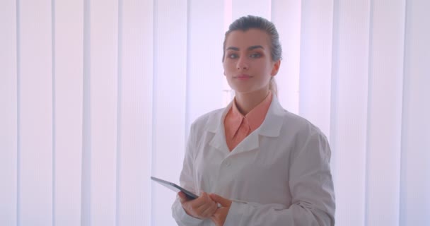 Närbild porträtt av unga attraktiva kaukasiska kvinnlig läkare som innehar en tablett tittar på kameran stående inomhus — Stockvideo