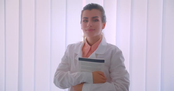 Closeup portrait of young attractive caucasian female doctor practitioner holding a book looking at camera standing indoors — Stock Video