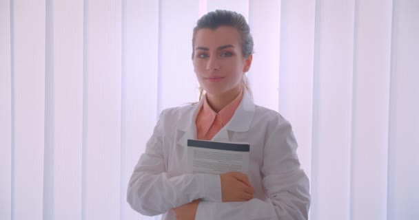 Крупный план портрета молодой привлекательной кавказской женщины-врача, держащей книгу, смотрящей в камеру, счастливо улыбающейся стоя в помещении — стоковое видео