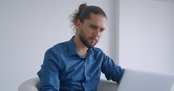 Modern freelancer with ponytail working with laptop sitting in armchair in light office turns to camera and laughing happily. — Stock Video