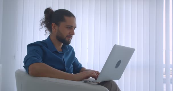 Photo de profil d'un pigiste moderne tapant sur un ordinateur portable assis dans un fauteuil dans un bureau léger se tourne vers la caméra et regarde avec sourire . — Video