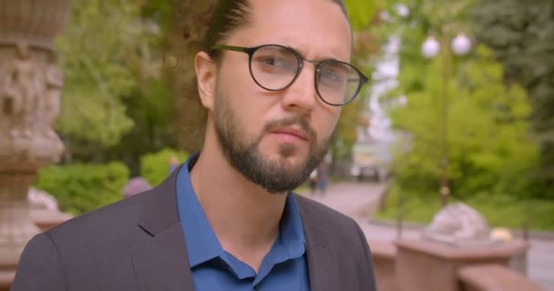 Bel homme d'affaires hipster dans des lunettes avec queue de cheval regarder sérieusement dans la caméra près de la fontaine dans le parc . — Video