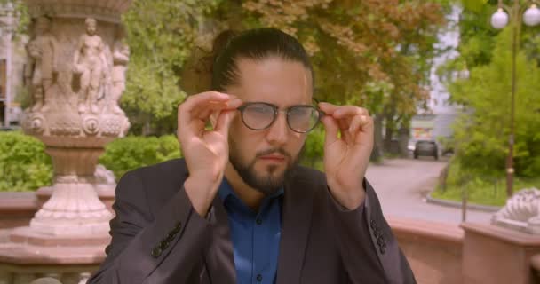 Handsome hipster businessman wears eyeglasses with ponytail and smiles into camera being positive in park. — Stock Video