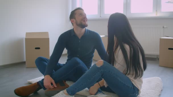 Unga kaukasiska par sitter på golvet i nya huset nära fönster och cardboxes drömmer om livet i nya hem. — Stockvideo