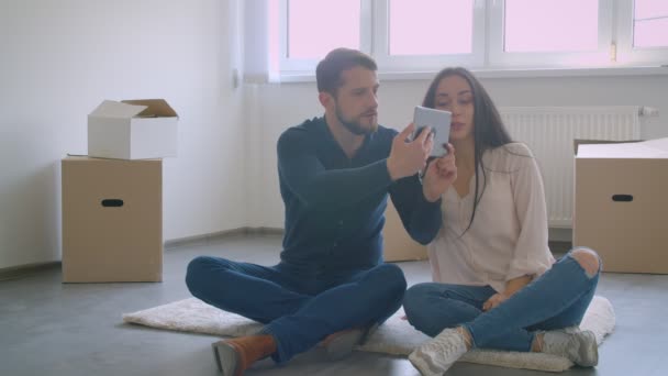 カートンボックスの近くの床に座っている間、デジタルタブレットを使用して話し、笑顔を使用して美しい白人のカップル. — ストック動画