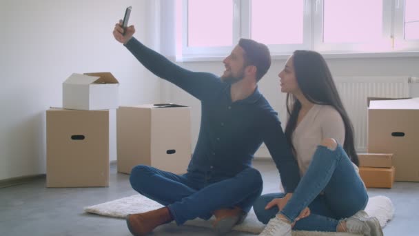 Bella coppia caucasica fare selfie utilizzando smartphone mentre seduto sul pavimento vicino scatole di cartone . — Video Stock
