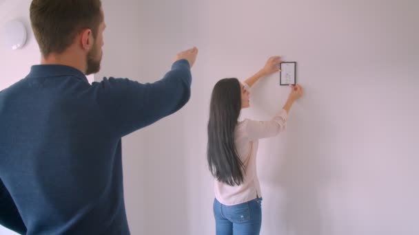 Caucásico chica morena colgando imagen enmarcada en la pared blanca, mientras que su novio ordena el lugar adecuado para ello . — Vídeo de stock