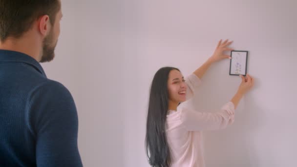 Kaukasisches fröhliches Mädchen hängt gerahmtes Bild an weiße Wand, während ihr Freund sie an die richtige Stelle dirigiert. — Stockvideo