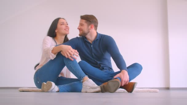 Fiatal kaukázusi házaspár ül a padlón az új üres lakás átölelve és megcsókolta, hogy boldog a fehér fal háttér — Stock videók